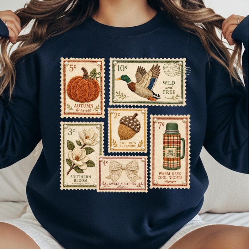 May include: Navy blue sweatshirt featuring vintage-style postage stamp designs. Stamps depict a pumpkin, duck, acorn, magnolia, bow, and a thermos. Text includes "Autumn Harvest," "Wild and Free," "Nature's Treasure," "Southern Bloom," "Sweet Southern Charm," and "Warm Days Cool Nights."