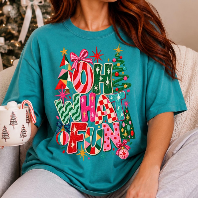 K&ouml;nnte beinhalten: Ein blaugr&uuml;nes Kurzarm-T-Shirt mit einer bunten Grafik und dem Schriftzug "OH WHAT FUN" in verschiedenen Mustern und Farben. Das Design zeigt stilisierte Weihnachtsb&auml;ume, rosa Schleifen und Christbaumkugeln, umgeben von Sternen.