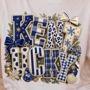 May include: A white t-shirt featuring a large, decorative graphic that spells out KENTUCKY. The letters are filled with various blue and white patterns, including plaid, leopard print, polka dots, and snowflakes, accented by bows and ornaments.
