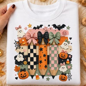 K&ouml;nnte beinhalten: Wei&szlig;es Sweatshirt mit einem Halloween-Motiv. Das Design zeigt drei Bleistifte mit Schleifen, Geistern, K&uuml;rbissen, Flederm&auml;usen, Sternen und S&uuml;&szlig;igkeiten. Die Bleistifte haben karierte und Sternmuster.