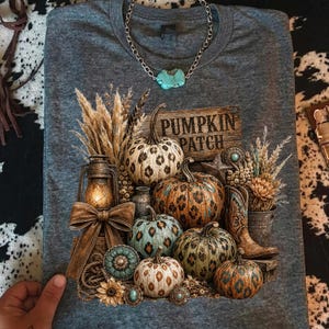 May include: Gray t-shirt featuring a fall-themed design with pumpkins, wheat, a lantern, and a sign that reads "Pumpkin Patch." The pumpkins have various patterns and colors. A turquoise necklace is draped on the shirt.