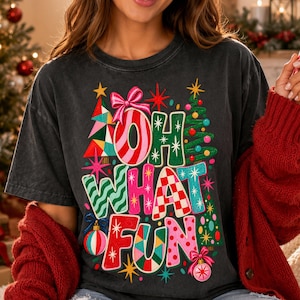 K&ouml;nnte beinhalten: Ein anthrazitfarbenes Kurzarm-T-Shirt mit einer bunten Weihnachtsgrafik und dem Schriftzug OH WHAT FUN. Das Design zeigt stilisierte Weihnachtsb&auml;ume, rosa Schleifen, Christbaumkugeln und Sternmotive in den Farben Rot, Gr&uuml;n, Pink und Blau.