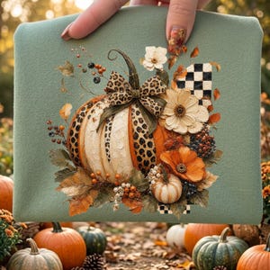 May include: A sage green fabric featuring a central graphic of a pumpkin adorned with a leopard print bow, surrounded by orange and white flowers, berries, autumn leaves, and a black and white checkered pattern element.