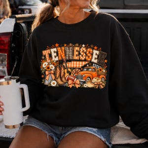 Może przedstawiać: Czarna bluza z okrągłym dekoltem z nadrukiem graficznym, kt&oacute;ry układa się w napis TENNESSEE wzorzystymi literami blokowymi. Projekt zawiera gitarę, kowbojki, piłkę do futbolu amerykańskiego, pomarańczowy pickup, kowbojski kapelusz i słoneczniki.