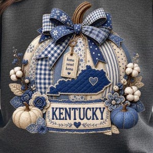 May include: A gray sweatshirt featuring a decorative pumpkin design. The pumpkin is adorned with blue and white patterned fabric, a large bow, and the word "Kentucky." Additional elements include a tag that says "home sweet home."