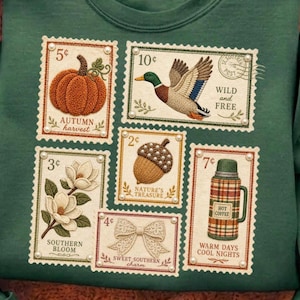 Puede incluir: Sudadera verde con diseños de sellos postales con temas otoñales y sureños. Los sellos representan una calabaza, un pato, una bellota, una magnolia, un lazo y un termo. Textos: "Autumn Harvest", "Wild and Free", "Nature's Treasure", "Southern Bloom", "Sweet Southern Charm" y "Warm Days Cool Nights."