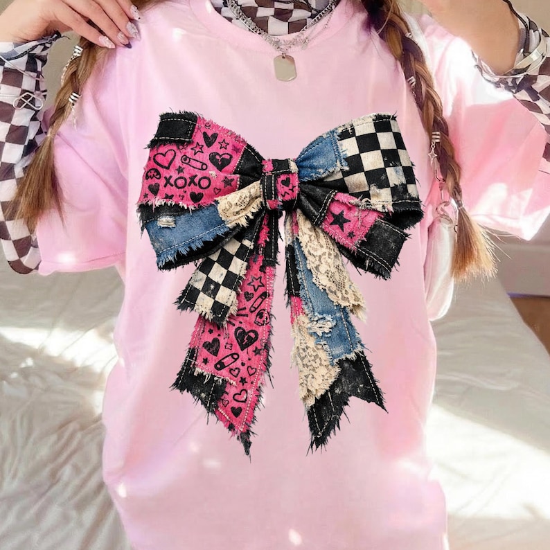 Op de afbeelding: Roze T-shirt met een groot patchwork strikdesign. De strik bevat denim, zwart-wit geruite stof en roze stof met harten, sterren en de tekst "XOXO". De strik heeft een gerafelde rand.