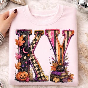 Pode incluir: Uma camisola de gola redonda rosa claro com um gr&aacute;fico grande das letras KY. A letra K est&aacute; decorada com um c&eacute;u noturno, estrelas, uma lua crescente, folhas de outono, uma ab&oacute;bora e uma vela. A letra Y apresenta riscas, livros e um caldeir&atilde;o.
