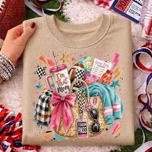 May include: A tan crewneck sweatshirt featuring a graphic print of a straw tote bag filled with a tumbler labeled I'm That Mom, a football, a game schedule, a pink bow, a plaid scarf, a teal sweater, sunglasses, and a Stadium Pass tag.