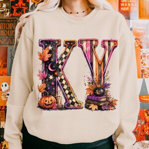 Pode incluir: Uma camisola bege de gola redonda com um grande gr&aacute;fico das letras KV. O K est&aacute; decorado com um padr&atilde;o axadrezado preto e branco, estrelas, uma lua crescente, uma vela acesa e uma ab&oacute;bora. O V apresenta riscas cor de laranja e pretas.