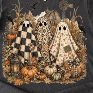 May include: A charcoal grey t-shirt featuring a graphic print of three ghosts draped in patterned fabrics, including black and white buffalo check, leopard print, and floral. The scene includes pumpkins, a lantern, crows, and a barn.