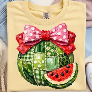 May include: Pale yellow t-shirt with a watermelon design. The watermelon is decorated with a patchwork pattern, a pink polka-dot bow, and a red bow. A slice of watermelon is next to the main design. The shirt has a crew neck.