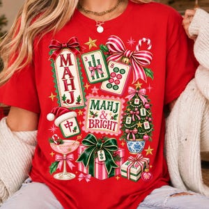 K&ouml;nnte beinhalten: Ein leuchtend rotes T-Shirt mit einer festlichen Grafik aus Mahjong-Steinen, einem mit Mahjong-Steinen geschm&uuml;ckten Weihnachtsbaum, Geschenken, einem Cocktailglas und Schleifen. Der Text lautet "MAHJ" und "MAHJ & BRIGHT" in Rot und Gr&uuml;n.