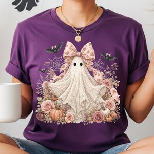 Pode incluir: Uma t-shirt roxa de manga curta com um gr&aacute;fico de um fantasma branco a usar um la&ccedil;o grande com padr&atilde;o, rodeado por rosas cor-de-rosa, pequenas ab&oacute;boras, teias de aranha e pequenos morcegos pretos. O fantasma tem dois olhos pretos.