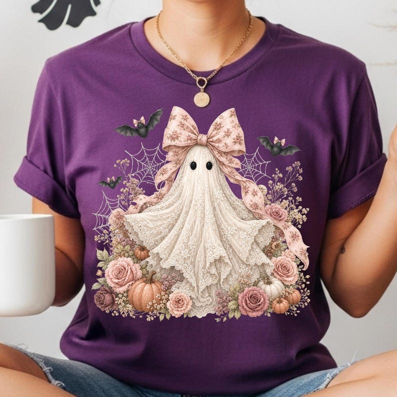 Pode incluir: Uma t-shirt roxa de manga curta com um gr&aacute;fico de um fantasma branco a usar um la&ccedil;o grande com padr&atilde;o, rodeado por rosas cor-de-rosa, pequenas ab&oacute;boras, teias de aranha e pequenos morcegos pretos. O fantasma tem dois olhos pretos.