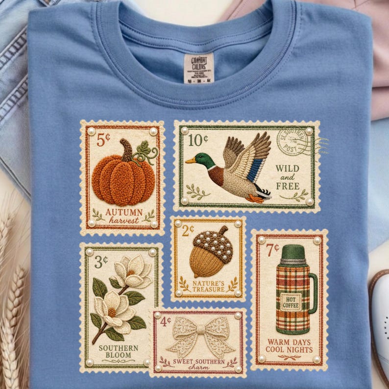May include: Light blue t-shirt featuring postage stamp designs with autumn and southern themes. Stamps depict a pumpkin, duck, acorn, magnolia, bow, and a thermos. Text includes "Autumn Harvest", "Wild and Free", "Nature's Treasure", "Southern Bloom", "Sweet Southern Charm", and "Hot Coffee".