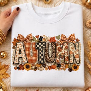 May include: A white crewneck sweatshirt featuring the word AUTUMN in large, patterned letters. The design includes plaid, leopard print, and floral motifs, accented with gingham bows, small buttons, sunflowers, and various fall leaves.