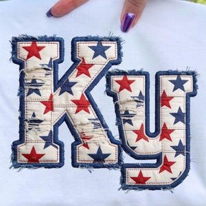 May include: White sweatshirt featuring the letters "KY" in a patriotic design. The letters are appliqued with a distressed, star-patterned fabric in red and blue, outlined with a frayed denim trim.