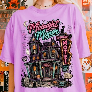 May include: A lilac-colored t-shirt featuring a spooky illustration of a haunted house with the text "The Midnight Manor." The design includes a motel sign, pumpkins, ghosts, and a black cat, perfect for Halloween.