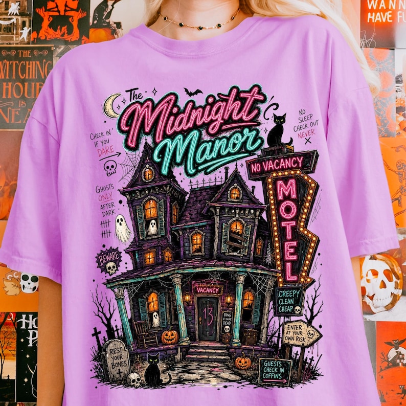 May include: A lilac-colored t-shirt featuring a spooky illustration of a haunted house with the text "The Midnight Manor." The design includes a motel sign, pumpkins, ghosts, and a black cat, perfect for Halloween.