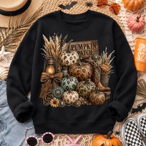 May include: Black sweatshirt featuring a fall-themed graphic. The design includes various pumpkins with leopard print patterns, a wooden sign that reads "Pumpkin Patch", and a lantern. The overall color palette includes shades of orange, brown, and teal.