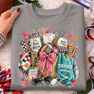 May include: A grey crewneck sweatshirt featuring a graphic print of a straw tote bag filled with a tumbler labeled 'I'm That Mom', a football, a leopard print cap, a teal sweater, a plaid blanket, sunglasses, and a stadium pass.