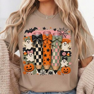 K&ouml;nnte beinhalten: Beiges T-Shirt mit einem Halloween-Motiv. Das Design zeigt drei Bleistifte mit Schleifen, Geistern, K&uuml;rbissen, Flederm&auml;usen und Spinnweben. Die Bleistifte sind schwarz-wei&szlig; kariert, orange mit Sternen und rosa und gr&uuml;n kariert.