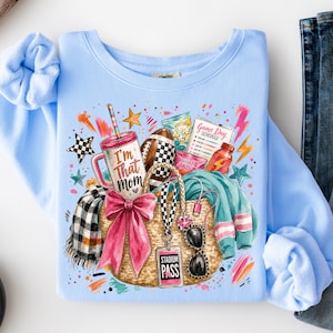 May include: A light blue crewneck sweatshirt featuring a graphic print of a straw tote bag with a large pink bow, a tumbler labeled I'm That Mom, a football, a stadium pass, sunglasses, a plaid scarf, and a game day schedule with colorful accents.