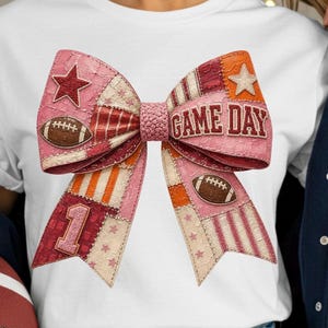 May include: A pink and patchwork bow with the words "GAME DAY" in the center. The bow features footballs, stars, and the number 1. The bow is on a white t-shirt.