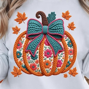 Puede incluir: Una sudadera blanca con un gráfico central de una calabaza con un exterior de punto naranja texturizado y un gran lazo de ganchillo azul turquesa en la parte superior. Los paneles de la calabaza tienen bordados florales y hojas de arce naranjas.