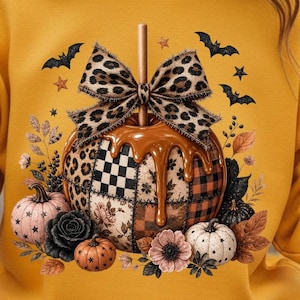 May include: Mustard yellow sweatshirt featuring a caramel apple design with a leopard print bow, surrounded by pumpkins, bats, and stars. The apple has a patchwork design with floral, plaid, and checkered patterns, perfect for fall and Halloween.