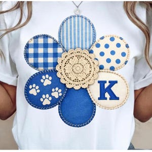 May include: White t-shirt with a large, flower-shaped applique. The applique features blue and white patterned circles, a beige lace center, and a circle with the letter "K". The design includes paw prints and gingham patterns.