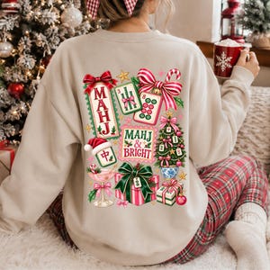 K&ouml;nnte beinhalten: Ein beigefarbenes Sweatshirt mit Rundhalsausschnitt und einem farbenfrohen R&uuml;ckendruck aus Mahjong-Steinen, einem Weihnachtsbaum, einem Geschenk und einem Cocktailglas. Der Text lautet "MAHJ" und "MAHJ & BRIGHT" mit roten und gr&uuml;nen Schleifen.