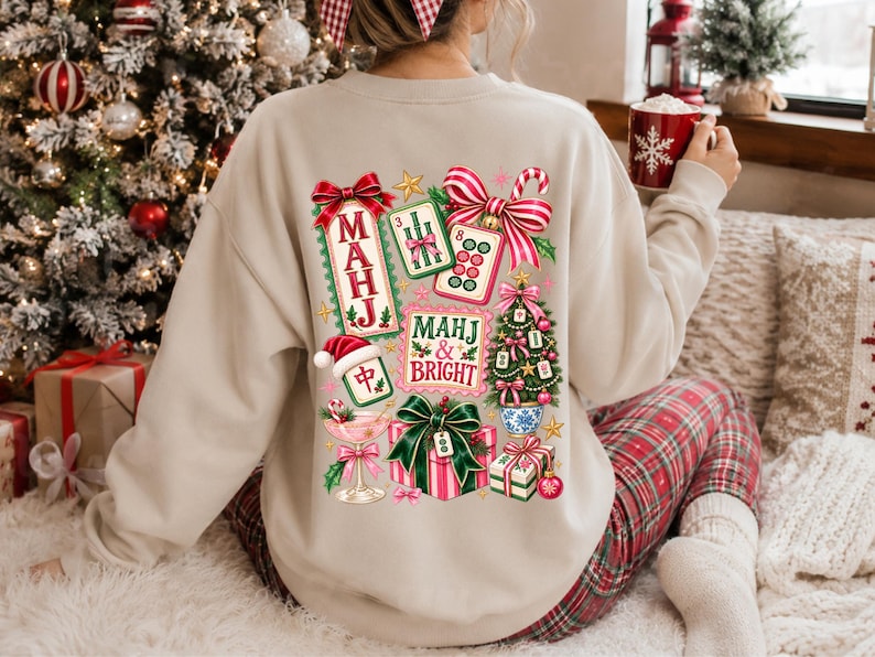K&ouml;nnte beinhalten: Ein beigefarbenes Sweatshirt mit Rundhalsausschnitt und einem farbenfrohen R&uuml;ckendruck aus Mahjong-Steinen, einem Weihnachtsbaum, einem Geschenk und einem Cocktailglas. Der Text lautet "MAHJ" und "MAHJ & BRIGHT" mit roten und gr&uuml;nen Schleifen.