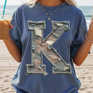 May include: A blue t-shirt featuring a large, distressed letter "K" applique. The letter is outlined with a silver wire-like material and has a denim-like texture with patches of white and red fabric peeking through.