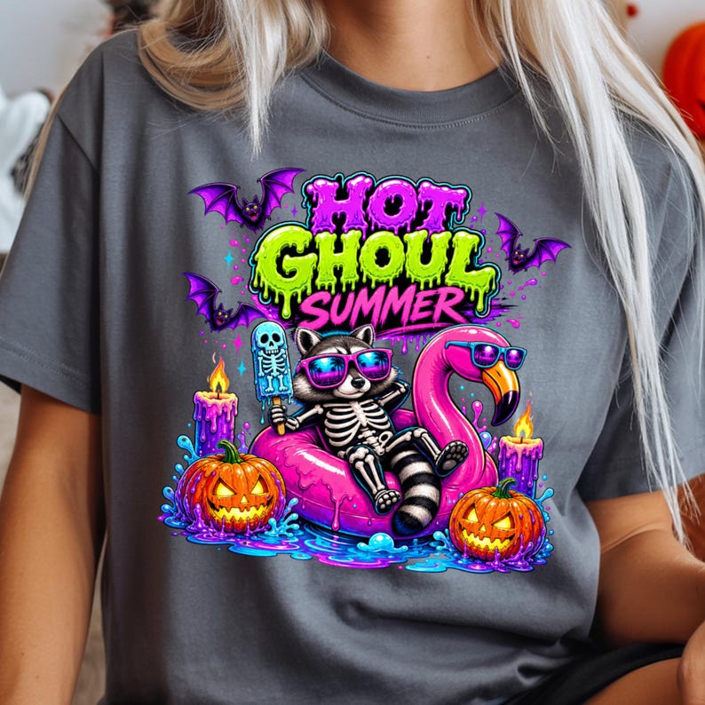 May include: Gray t-shirt with a colorful graphic featuring a skeleton raccoon wearing sunglasses, lounging on a pink flamingo float. The design includes pumpkins, candles, bats, and the text "HOT GHOUL SUMMER."