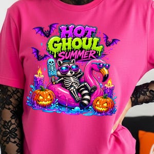 May include: Pink t-shirt with a Halloween-themed graphic. The design features a raccoon skeleton wearing sunglasses, floating on a pink flamingo floatie, holding a skeleton popsicle. The text "HOT GHOUL SUMMER" is above the raccoon. Pumpkins, candles, and bats complete the design.