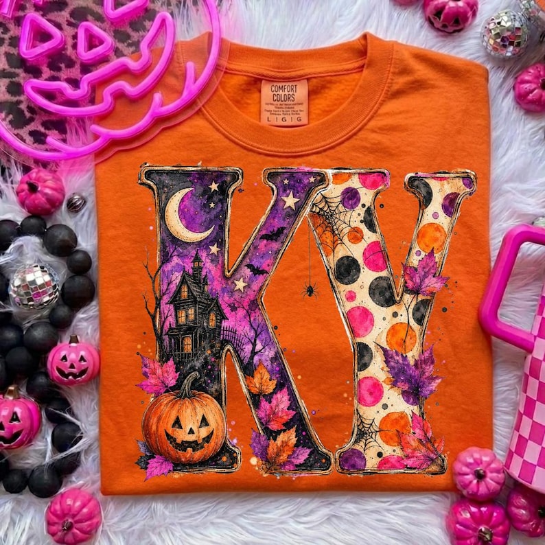 Pode incluir: Uma t-shirt laranja com um gr&aacute;fico grande das letras KV. O K est&aacute; preenchido com uma casa assombrada, uma lua crescente e uma ab&oacute;bora. O V apresenta um padr&atilde;o de bolinhas, teias de aranha e folhas de outono em tons roxos e laranja.