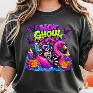 May include: Dark gray t-shirt with a colorful graphic. The graphic features a skeleton raccoon wearing sunglasses and floating on a pink flamingo pool float. The text "HOT GHOUL SUMMER" is above the raccoon, with bats, pumpkins, and candles.