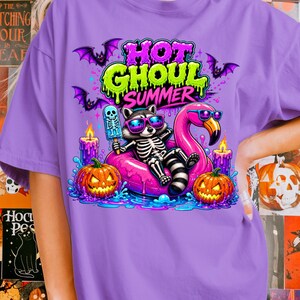 May include: A purple t-shirt with a colorful graphic featuring a skeleton raccoon wearing sunglasses, floating on a pink flamingo pool float, holding a popsicle. The text "HOT GHOUL SUMMER" is above the raccoon, with bats and jack-o'-lanterns.