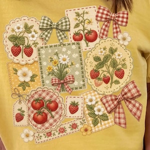 Può includere: T-shirt giallo con un design patchwork di fragole, fiori e fiocchi. Le toppe presentano fragole, pomodori e composizioni floreali. I fiocchi hanno un motivo a quadretti rossi e verdi.