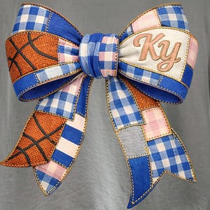 Könnte beinhalten: Eine große dekorative Schleife mit einem Patchwork-Design aus blau-weißem Vichy-Karo, orangefarbenem, basketball-texturiertem Stoff mit schwarzen Linien und einem cremefarbenen Abschnitt mit den gestickten Buchstaben Ky in Schreibschrift auf einem grauen Shirt.