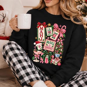 K&ouml;nnte beinhalten: Ein schwarzes Langarm-Sweatshirt mit einem bunten Grafikdruck aus Mahjong-Steinen, einem Weihnachtsbaum, verpackten Geschenken, einer Zuckerstange und einem Cocktailglas. Der Text lautet MAHJ und MAHJ & BRIGHT in festlichen roten und gr&uuml;nen Buchstaben.
