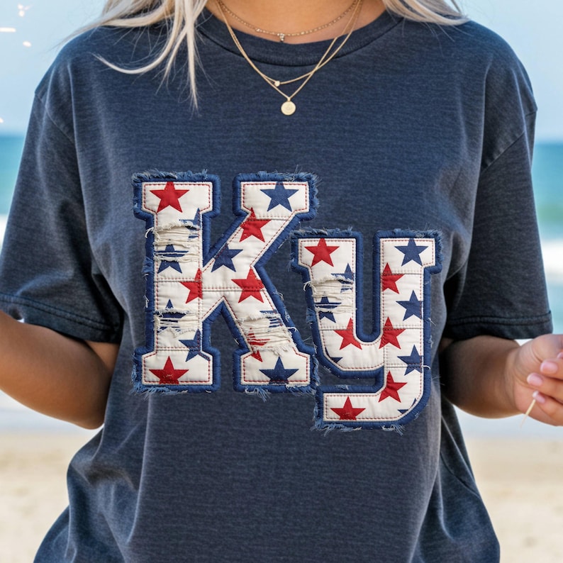 May include: Navy blue t-shirt with the letters "KY" appliqued on the front. The letters are filled with a pattern of red and blue stars on a white background, with a distressed denim border.