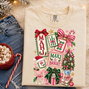 K&ouml;nnte beinhalten: Ein cremefarbenes T-Shirt mit einem festlichen Grafikdruck aus Mahjong-Steinen, einem Weihnachtsbaum, verpackten Geschenken, einem Cocktailglas und Zuckerstangen. Der Text lautet "MAHJ" und "MAHJ & BRIGHT" in roter und gr&uuml;ner Schrift mit Feiertagsmotiven.