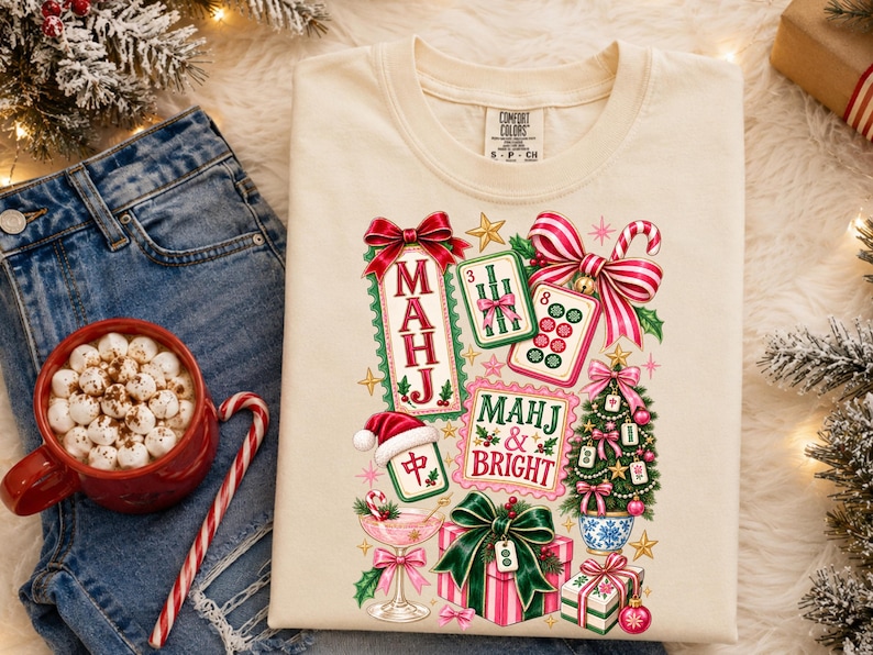 K&ouml;nnte beinhalten: Ein cremefarbenes T-Shirt mit einem festlichen Grafikdruck aus Mahjong-Steinen, einem Weihnachtsbaum, verpackten Geschenken, einem Cocktailglas und Zuckerstangen. Der Text lautet "MAHJ" und "MAHJ & BRIGHT" in roter und gr&uuml;ner Schrift mit Feiertagsmotiven.