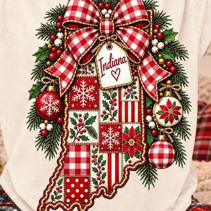 May include: A cream-colored t-shirt featuring a graphic of the state of Indiana in a patchwork quilt design. The graphic includes a red and white gingham bow, pine branches, ornaments, and a tag with the text Indiana and a heart symbol.