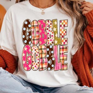 May include: A white short-sleeved t-shirt featuring the text GOBBLE GOBBLE in large, blocky letters. Each letter is filled with a different colorful pattern, including polka dots, plaid, and stars in shades of brown, pink, orange, and green.