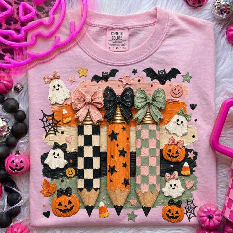 K&ouml;nnte beinhalten: Rosa Comfort Colors Sweatshirt mit einem Halloween-Design. Das Design zeigt drei Bleistifte mit Schleifen, Geistern, K&uuml;rbissen, Flederm&auml;usen und Sternen. Die Bleistifte haben karierte und Sternmuster.