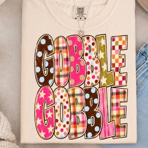 May include: A cream-colored t-shirt featuring the text GOBBLE GOBBLE in two rows. Each letter is filled with a different pattern, including brown with blue dots, plaid, solid pink, multi-colored polka dots, olive green with stars, and orange checks.